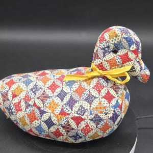May include: A stuffed fabric duck featuring a colorful patchwork pattern of red, blue, yellow, and white geometric shapes. The duck has a small black bead eye and a bright yellow ribbon tied in a bow around its neck, creating a decorative accent.