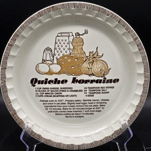 May include: A round ceramic pie dish with a fluted edge and brown rim detailing. The center features a gold-toned illustration of kitchen tools, cheese, and onions, with the text Quiche Lorraine and a printed recipe in black ink.