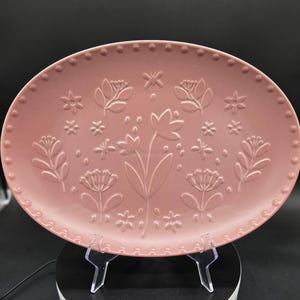 May include: A dusty rose oval serving platter with embossed floral designs. The platter features a border of small raised dots and various flower and leaf patterns. The serving dish is displayed on a clear plastic stand against a dark background.