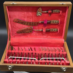 May include: A wooden storage box with a red velvet interior containing a set of gold-toned metal flatware. The set includes a serving spoon, a large knife, a cake server, and multiple smaller forks, all featuring dark brown handles with embossed details.
