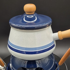 May include: A white enamel fondue pot with blue speckled details, featuring a dark blue lid with a light wood knob. The pot has a long light wood handle and sits on a dark blue metal stand. Two fondue forks with matching light wood handles are visible.