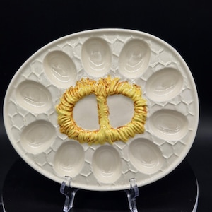 May include: Oval, off-white ceramic deviled egg serving platter with 12 egg indentations and a central pretzel-shaped design in yellow and brown. The platter has a textured, honeycomb-like pattern.