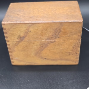 May include: A small, rectangular wooden box with a lid. The box is constructed from light-colored wood with visible grain and is assembled with dovetail joints at the corners. The box is sitting on a black surface.