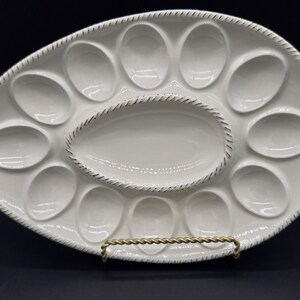 May include: An oval-shaped white ceramic deviled egg serving platter featuring twelve recessed egg-shaped indentations surrounding a central oval compartment. The outer rim and the central compartment edge are detailed with a textured rope-like pattern.
