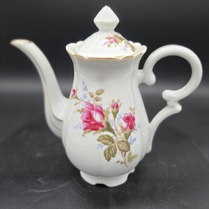 May include: White porcelain teapot with a floral rose design in pink, red, and green. The teapot has a curved spout, a loop handle, and a lid with a decorative finial. Gold trim accents the edges of the teapot and lid.