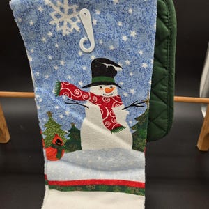 May include: A kitchen towel featuring a snowman wearing a black top hat and a red patterned scarf, set against a blue background with white snowflakes, green pine trees, and a red cardinal, paired with a dark green quilted oven mitt.