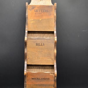 May include: Wooden wall-mounted organizer with three compartments labeled "LETTERS", "BILLS", and "MISCELLANEOUS". The top features a carved rooster design. The organizer is made of stained wood.