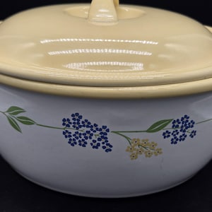 May include: A white enamel casserole dish with a matching pale yellow lid. The exterior features a floral pattern of small blue and yellow flowers with green leaves encircling the bowl. The pot has a smooth, glossy finish and a rounded shape.