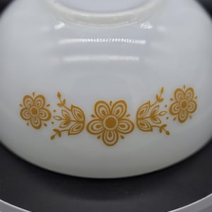 May include: A white milk glass bowl featuring a repeating horizontal pattern of gold-colored flowers and leaves. The design consists of three stylized blossoms connected by delicate leafy stems, printed directly onto the smooth, curved surface.