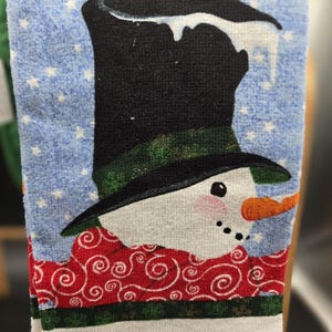 May include: A white kitchen towel featuring a printed illustration of a snowman wearing a black top hat with a green band, a red scarf with white swirl patterns, and an orange carrot nose against a light blue background with white stars.