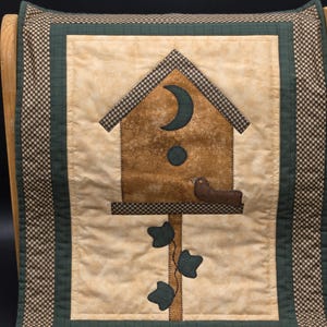 May include: A quilted wall hanging featuring a birdhouse design. The birdhouse is brown with a crescent moon and a small bird. The background is beige with a green border and a checkered pattern.