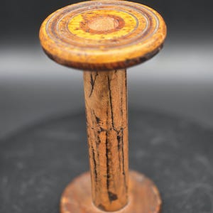 May include: A vintage wooden spool with a circular top and base, connected by a cylindrical stem. The wood has a warm, aged appearance with visible grain and dark streaks. The top features concentric circles in shades of brown and yellow.