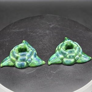 May include: Two ceramic candle holders in a floral design. The candle holders are glazed in shades of green and blue. The holders have a round opening for a candle and three leaf-shaped petals.