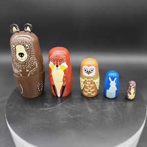 May include: A set of five wooden nesting dolls, each painted with a different animal: a bear, fox, owl, rabbit, and bird. The bear is brown, the fox is red, the owl is brown and orange, the rabbit is blue, and the bird is purple and yellow.