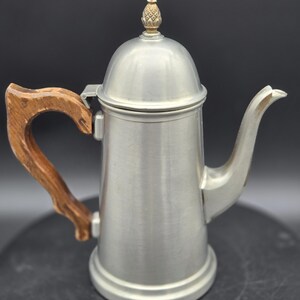May include: A silver-toned metal coffee pot with a smooth, cylindrical body and a curved spout. It features a brown, textured wooden handle and a domed lid topped with a small, decorative metallic finial shaped like a pineapple.