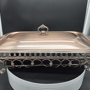 May include: A rectangular copper-toned chafing dish with a matching lid featuring a central handle. The base is supported by an ornate, open-work metal frame with curved detailing and four small feet, including side handles for easy transport.