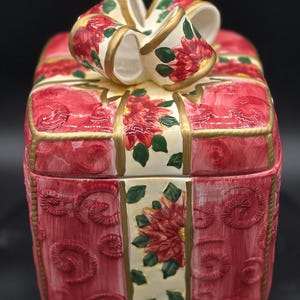 May include: A ceramic gift box container with a removable lid featuring a sculpted bow. The box is painted in shades of red with embossed swirl patterns and cream-colored bands decorated with red poinsettia flowers and green leaves, accented by gold trim.