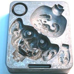 May include: A vintage metal mold with multiple cavities, including a circular shape and several detailed monster-like faces. The mold is marked with "©1985 MATTEL INC." and "4482-052A". The overall color is silver.