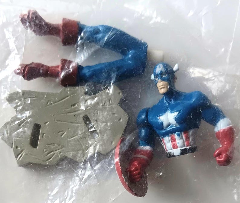 May include: A Captain America action figure in a plastic bag. The figure is blue, red, and white, with a shield and a star on the chest. The figure's legs are detached. The figure is a collectible toy.