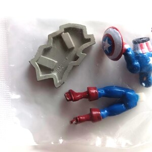 May include: A disassembled Captain America action figure with a shield, legs, and torso. The figure is primarily blue, red, and white, with a silver mold. The shield has a white star on a blue and red background. The pieces are on a white surface.