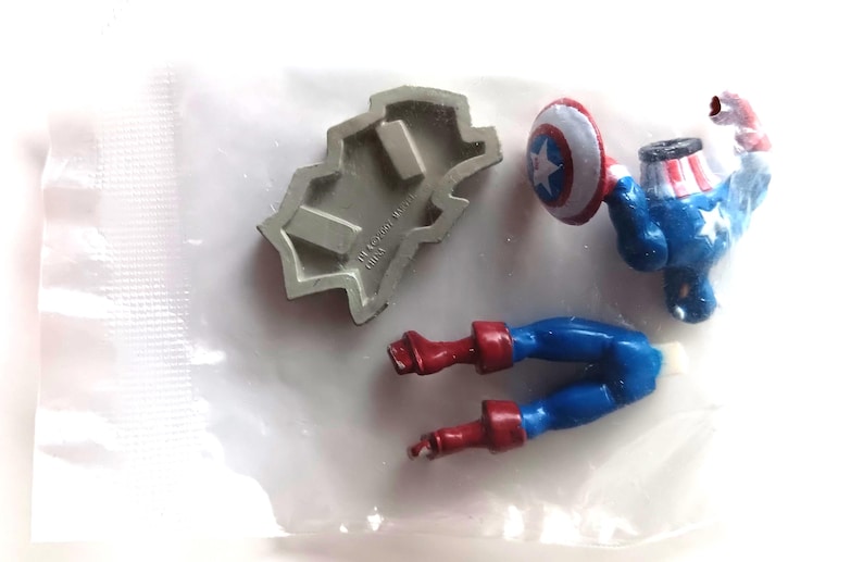 May include: A disassembled Captain America action figure with a shield, legs, and torso. The figure is primarily blue, red, and white, with a silver mold. The shield has a white star on a blue and red background. The pieces are on a white surface.