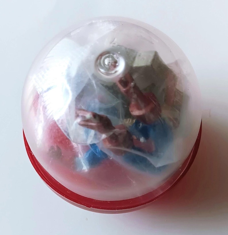 May include: A red and clear plastic capsule containing a small action figure. The figure is partially visible, with red, blue, and flesh-toned details. The capsule has a red base and a clear, rounded top.