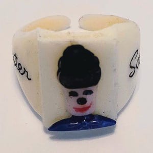 May include: A white ring with a cartoon face design. The face has a black hat, a red smile, and a blue collar. The word "Jen" is written on one side of the ring, and the word "So" is written on the other side.