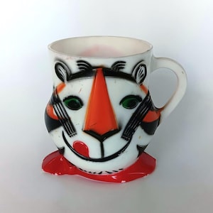 May include: A white ceramic mug shaped like a tiger face with a prominent orange nose, green eyes, and black stripes. The mug features a red tongue licking the lips and a red base. It has a curved handle on the right side and a smooth finish.