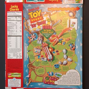 K&ouml;nnte beinhalten: Eine flachgedr&uuml;ckte Lucky Charms M&uuml;slipackung mit einer Toy Story Brettspielgrafik auf der R&uuml;ckseite. Das Design umfasst ein rotes, gelbes und blaues Farbschema mit Illustrationen von Spielzeug, einem Pfad und Textanweisungen f&uuml;r ein Wettrennen.