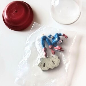 May include: A Captain America action figure in a plastic bag, with a red lid and a clear plastic dome. The figure is blue, red, and white, and is partially assembled. The base is gray.