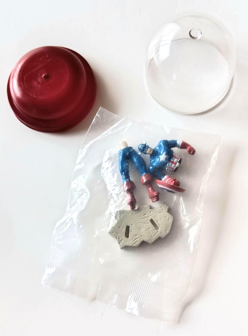 May include: A Captain America action figure in a plastic bag, with a red lid and a clear plastic dome. The figure is blue, red, and white, and is partially assembled. The base is gray.