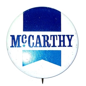May include: A round, white button with the name "McCarthy" in blue, with a blue star and two blue rectangular shapes. The button has a pin on the back.