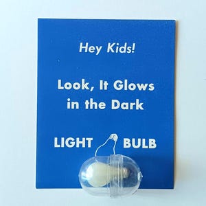 May include: A blue rectangular card with white text that reads Hey Kids! Look, It Glows in the Dark and LIGHT BULB. A small, clear plastic capsule containing a miniature white light bulb is attached to the bottom center of the blue card.