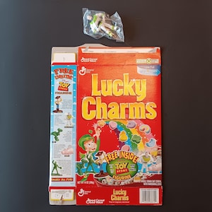 K&ouml;nnte beinhalten: Eine Lucky Charms M&uuml;slipackung mit Toy Story Branding, einem roten Hintergrund mit Regenbogen und Marshmallow-Formen sowie einer kleinen Buzz Lightyear Kunststofffigur in einer durchsichtigen T&uuml;te. Die Packung zeigt den Text Lucky Charms und Toy Story.