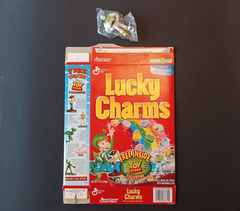 K&ouml;nnte beinhalten: Eine Lucky Charms M&uuml;slipackung mit Toy Story Branding, einem roten Hintergrund mit Regenbogen und Marshmallow-Formen sowie einer kleinen Buzz Lightyear Kunststofffigur in einer durchsichtigen T&uuml;te. Die Packung zeigt den Text Lucky Charms und Toy Story.