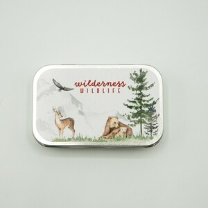 May include: A silver tin with a white background and a colorful illustration of a deer, a bear and a cub, and a pine tree. The text "wilderness WILDLIFE" is printed in red on the tin.