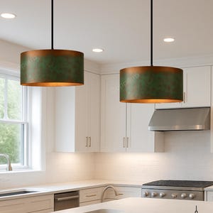 May include: Two pendant lights with cylindrical shades. The shades have a green and bronze finish. The lights hang from black cords in a modern kitchen. The kitchen features white cabinets and countertops.