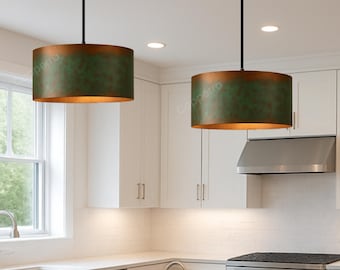 May include: Two pendant lights with cylindrical shades. The shades have a green and bronze finish. The lights hang from black cords in a modern kitchen setting. The kitchen features white cabinets and countertops.