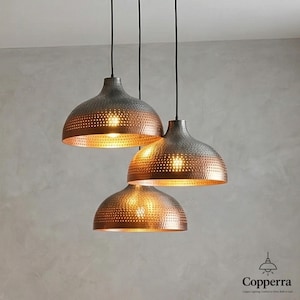 Peut inclure: Trois suspensions lumineuses en forme de dôme, avec un intérieur cuivré et un extérieur gris. Les lampes ont un design perforé et sont suspendues à des cordons noirs. L'image présente le logo "Copperra" en bas à droite.