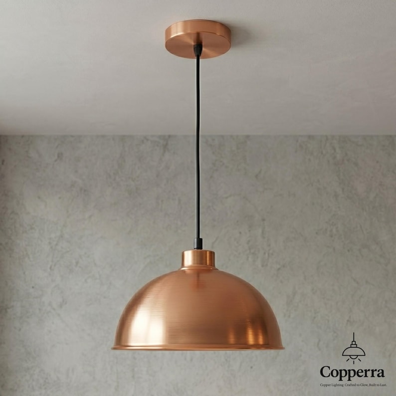 May include: A copper-colored pendant light fixture with a dome-shaped shade. The light hangs from a black cord attached to a circular ceiling mount. The fixture has a modern, industrial design, suitable for kitchen or dining room lighting.