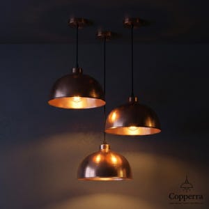 May include: Three copper pendant lights with a dome shape hang from the ceiling. The lights are illuminated, casting a warm glow. The fixtures have a metallic finish and are suspended by black cords.