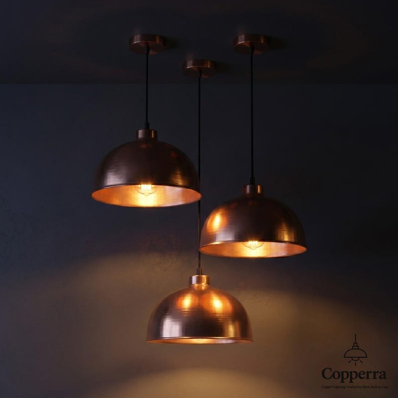 May include: Three copper pendant lights with a dome shape hang from the ceiling. The lights are illuminated, casting a warm glow. The fixtures have a metallic finish and are suspended by black cords.