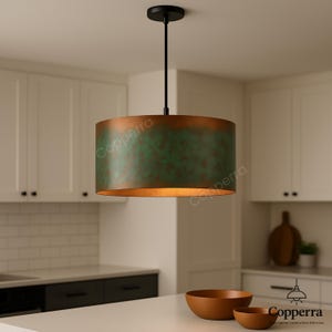 May include: A pendant light fixture with a cylindrical shade. The shade has a copper-colored exterior with a green patina and a gold interior. The fixture hangs from a black rod and canopy. The kitchen features white cabinets and wooden bowls.