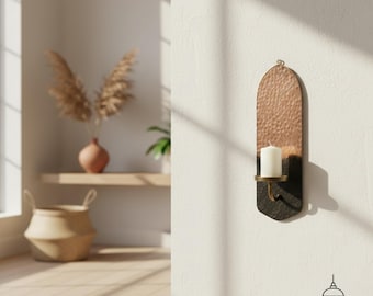 Puede incluir: Un portavelas de pared con la parte superior de cobre martillado y base negra. Una vela blanca se encuentra en un soporte de latón. El diseño es arqueado y tiene un lazo decorativo de metal en la parte superior para colgar.
