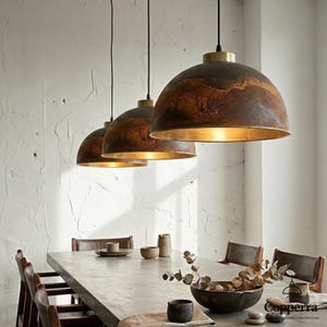 May include: Three pendant lights with a brown, textured exterior and a gold interior hang above a dining table. The lights have a dome shape and are suspended by black cords. The dining table is made of concrete and has a minimalist design.