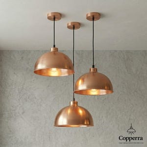 May include: Three copper-colored pendant lights hanging from the ceiling. Each light has a dome-shaped shade and a black cord. The lights are illuminated, casting a warm glow. The ceiling is white, and the wall is a textured gray.