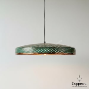 Puede incluir: Lámpara colgante verde y cobre. La pantalla circular está perforada con una textura martillada. La lámpara cuelga de un cable negro. La lámpara tiene una bombilla cálida.