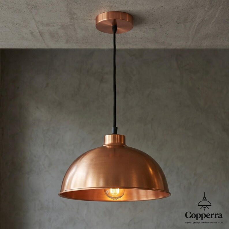 May include: A copper-colored pendant light fixture with a dome-shaped shade. The light hangs from a black cord and is attached to a circular ceiling mount. The light bulb is visible inside the shade. The fixture is a modern design.