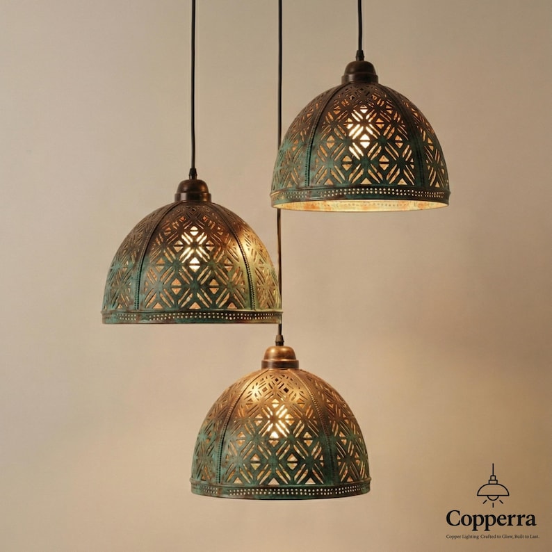 K&ouml;nnte beinhalten: Drei Pendelleuchten mit kuppelf&ouml;rmigen, gemusterten Schirmen in einem verwitterten T&uuml;rkis- und Bronzefinish. Die Leuchten h&auml;ngen an schwarzen Kabeln, und das Innere leuchtet mit warmem Licht. Das Logo "Copperra" ist sichtbar.