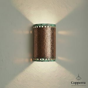 May include: A copper wall sconce with a textured surface and a verdigris rim. The cylindrical light fixture emits light upwards and downwards, creating a warm ambiance. The sconce is mounted on a light-colored wall.